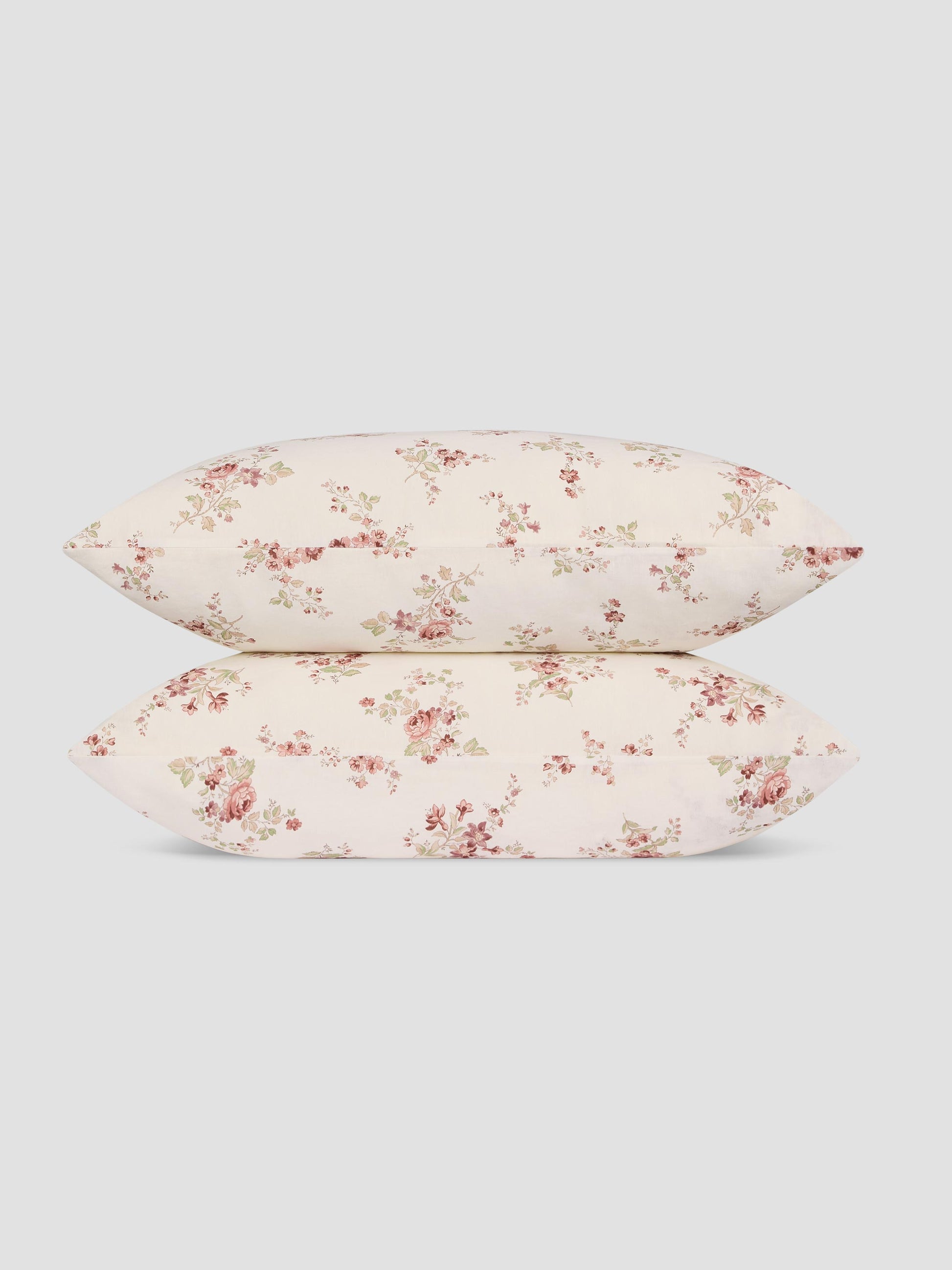 Two pillows stacked vertically, featuring a delicate pink and green floral pattern on a warm cream background, set against a solid light gray studio backdrop.