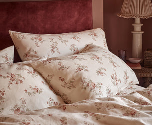 A close-up of a bed dressed in cream floral bedding with matching pillowcases and a dark red velvet headboard, set in a warmly lit bedroom with a bedside lamp.