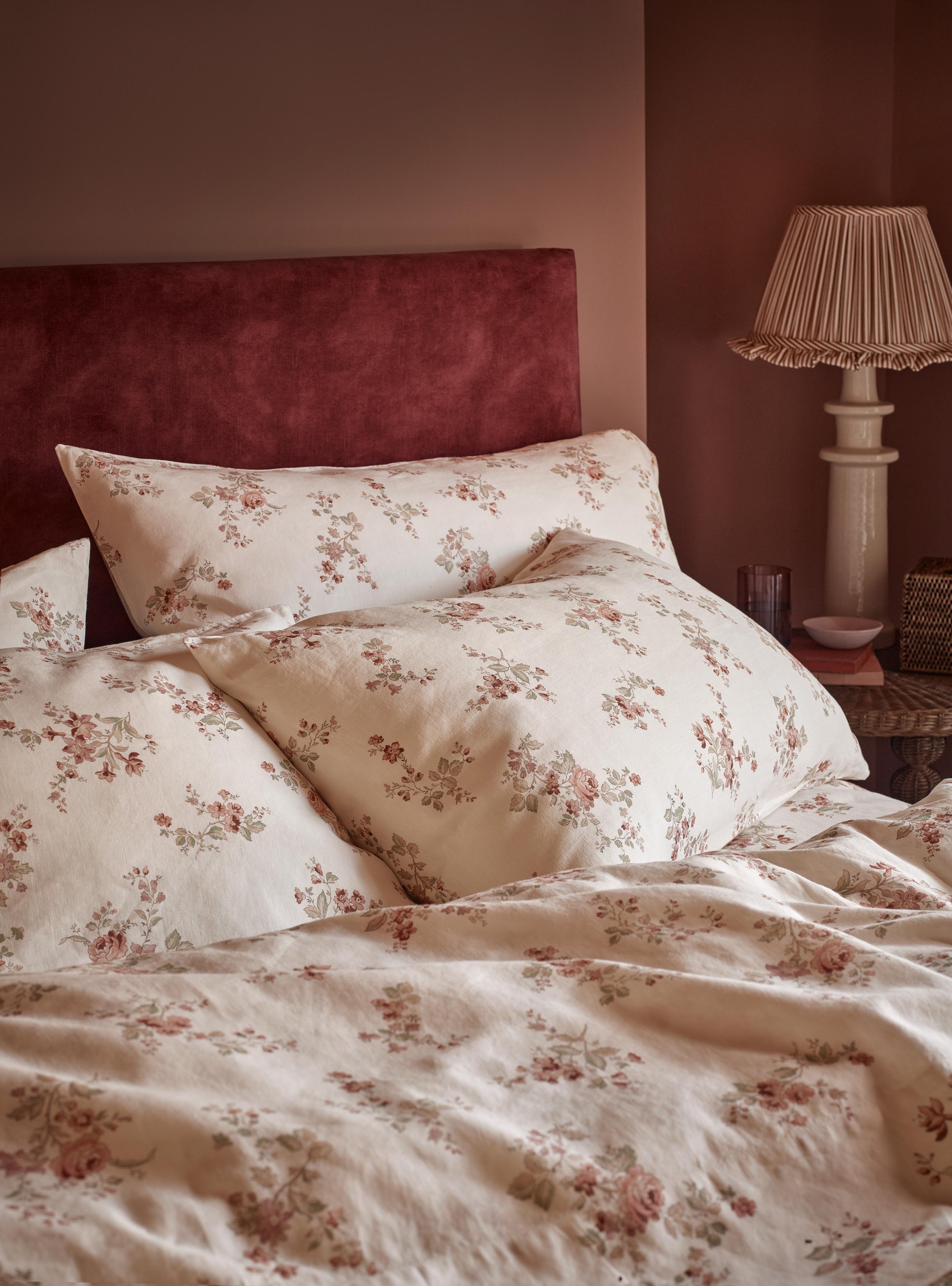 A close-up of a bed dressed in cream floral bedding with matching pillowcases and a dark red velvet headboard, set in a warmly lit bedroom with a bedside lamp.