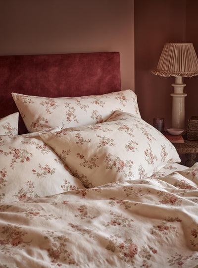 A close-up of a bed dressed in cream floral bedding with matching pillowcases and a dark red velvet headboard, set in a warmly lit bedroom with a bedside lamp.