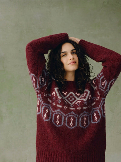 A woman with dark curly hair wearing a chunky knit burgundy sweater with a light blue and white geometric Fair Isle pattern across the chest and sleeves, posing with her arms raised against a textured sage green background.