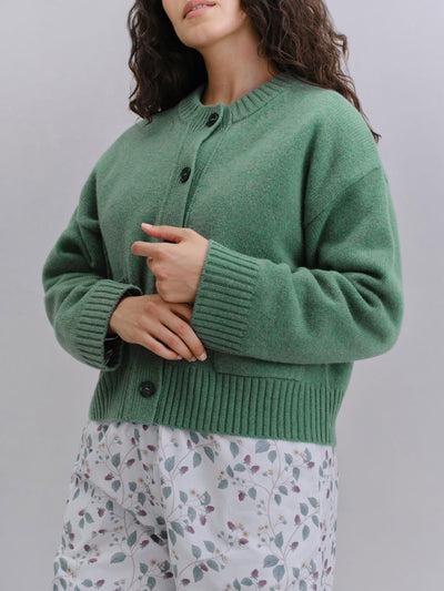 A woman wearing a sage green knit cardigan sweater with dark buttons, paired with white floral pajama pants, against a plain light gray background.