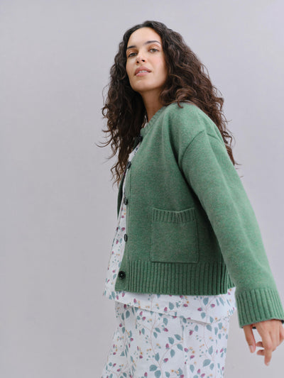 A woman wearing a sage green knit cardigan over a white floral pajama set, photographed against a solid light gray studio background.