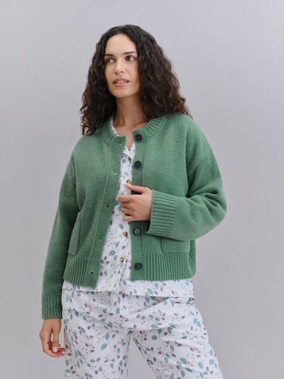 A woman modeling a sage green knit cardigan over a white floral pajama set, standing against a solid light gray studio background.