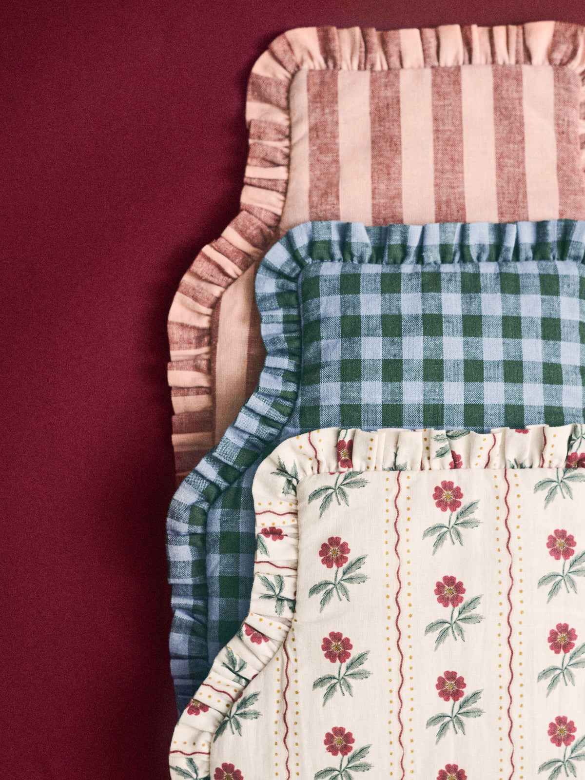 3 frill edged hot water bottles stacked on top of each other on a red background, one with a red floral pattern, one in dusk blue and fern green gingham and one rhubarb and rose pembroke stripe. 