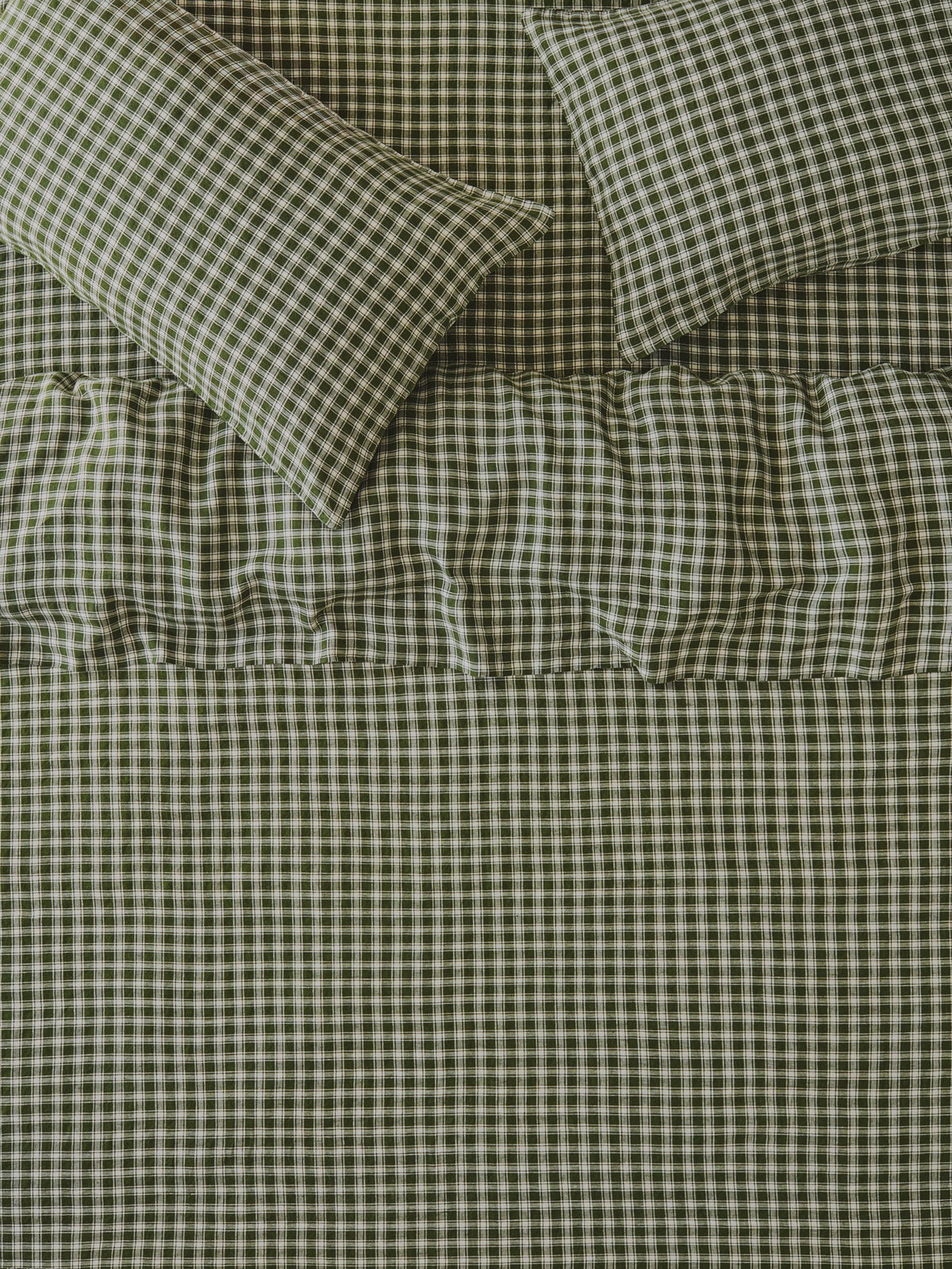 A close-up of a bed dressed in matching gingham bedding, showing a quilt or comforter and two pillows in an olive green and white check pattern with a ruffled band near the top.