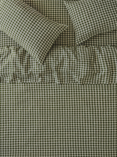 A close-up of a bed dressed in matching gingham bedding, showing a quilt or comforter and two pillows in an olive green and white check pattern with a ruffled band near the top.