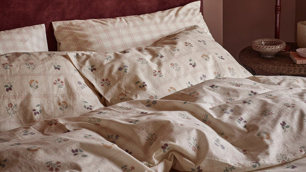 A close-up of a bed made with cream bedding featuring a delicate floral print and subtle plaid checks, styled in a warm bedroom setting.