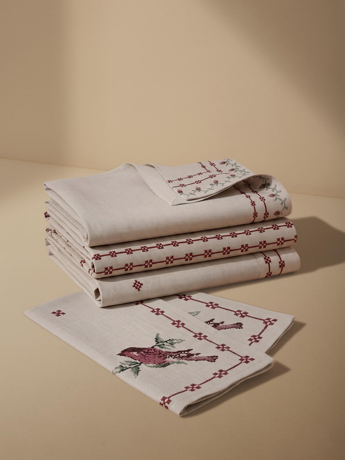 A stack of folded cream-colored linen table linens with red embroidered borders and a matching flat linen piece in front featuring a red bird and green leaf motif, set against a warm beige background.