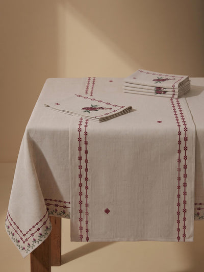 An off-white linen tablecloth with red embroidered borders and a matching stack of napkins on a wooden table against a warm beige background.