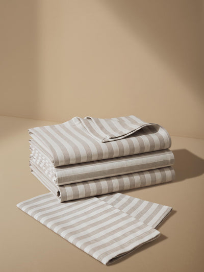 A stack of folded striped bedding or fabric pieces with alternating warm beige and light gray stripes, arranged on a matching beige surface against a beige backdrop.