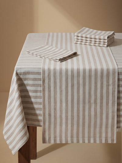 A beige and off-white vertically striped tablecloth draped over a wooden table, with matching folded napkins stacked on top, set against a warm beige studio background.