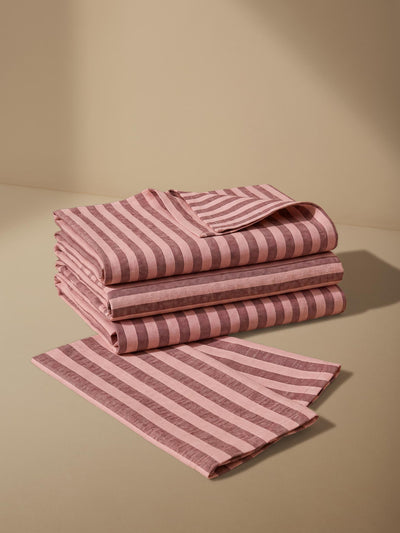 A stack of folded striped linens in dusty pink and muted mauve, with one matching striped cloth laid flat in front, set against a warm beige studio background.