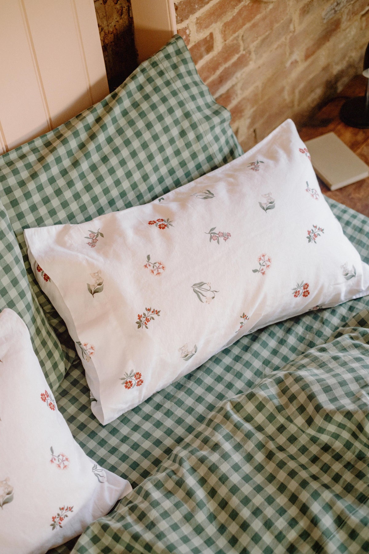 A white pillowcase with a delicate red and green floral print, styled on a bed with green and white gingham bedding and a brick wall background.