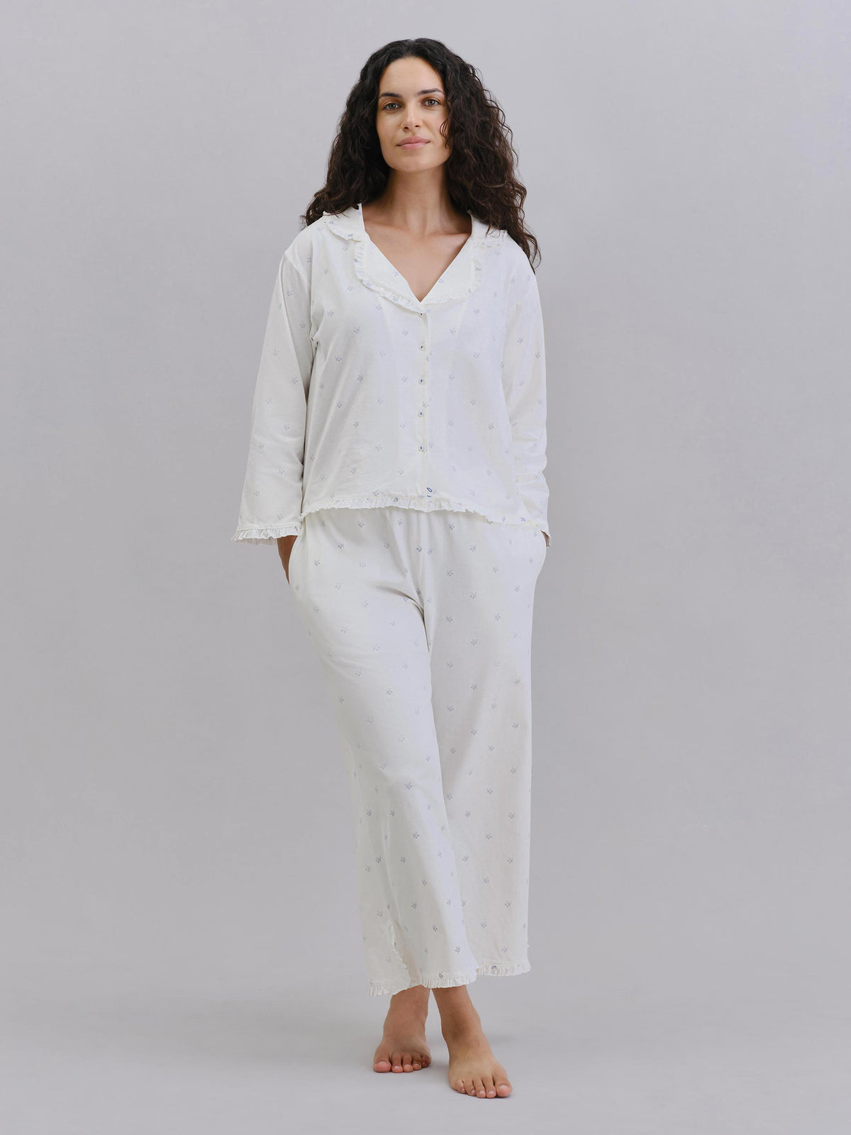 A woman modeling a two-piece white pajama set with a subtle dotted pattern, consisting of a long-sleeve button-front top and matching pants, against a solid gray studio background.
