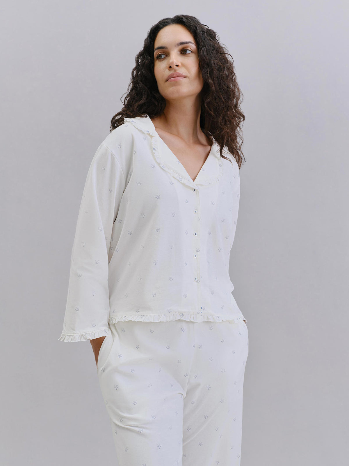 A woman modeling a white two-piece pajama set with a button-front long-sleeve top and matching pants, photographed against a plain light gray studio background.