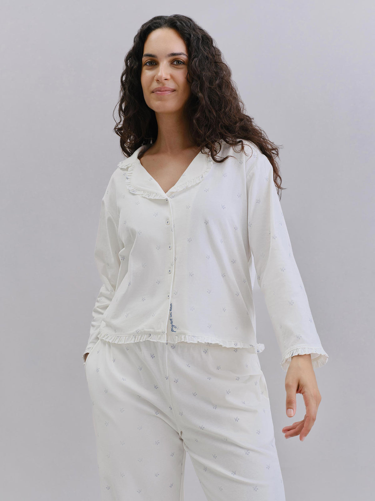 A woman modeling a white long-sleeve pajama set with a subtle dotted pattern, featuring a ruffled collar and button-front top paired with matching pants, against a plain light gray studio background.