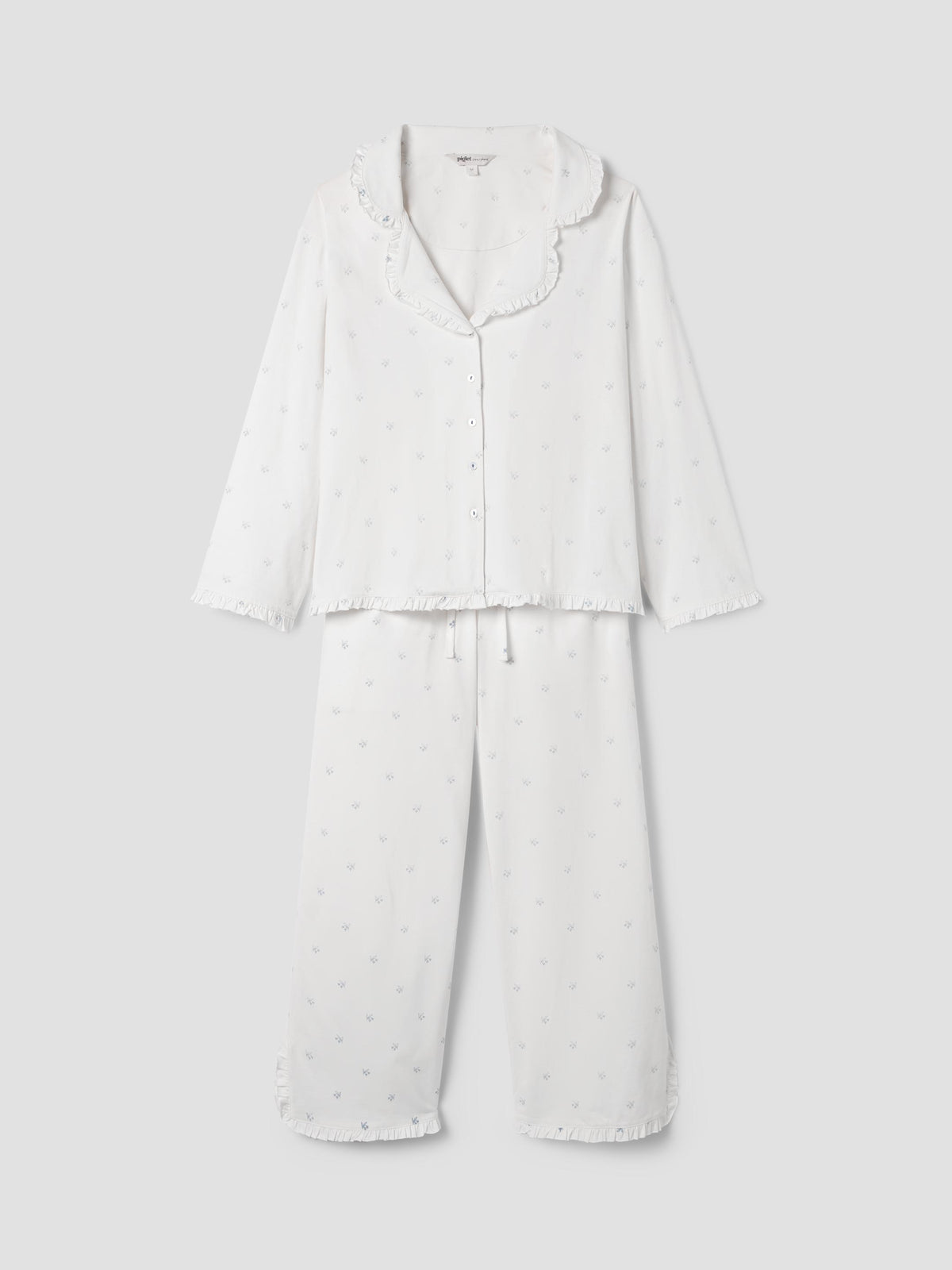 A two-piece white pajama set with a small polka dot pattern, featuring a long-sleeve button-up top with a ruffled collar and matching wide-leg pants, displayed on a neutral light gray background.