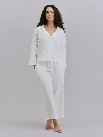 A woman modeling a two-piece white pajama set with a subtle dotted pattern, consisting of a long-sleeve button-front top and matching pants, against a solid gray studio background.