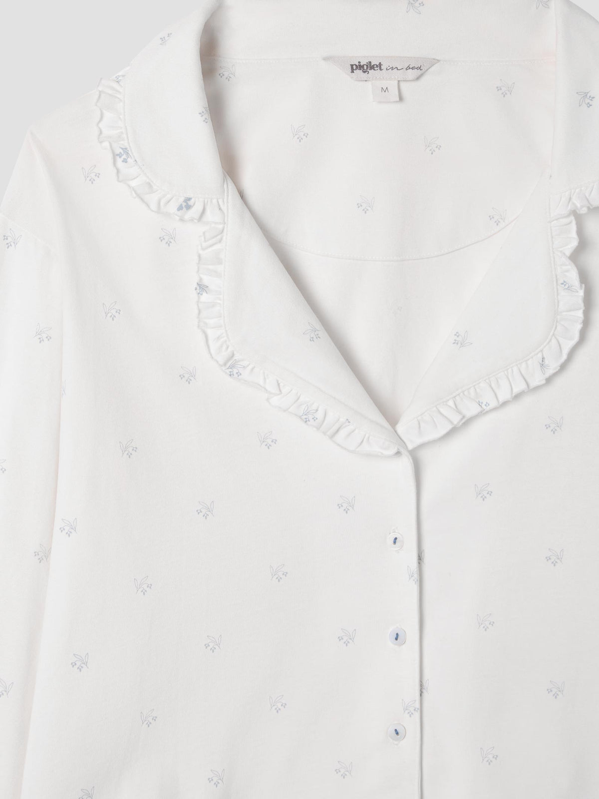 Close-up of a white long-sleeve pajama top with a ruffled Peter Pan collar and small scattered floral prints, showing the inner neck label with the brand 'petit luxe'.