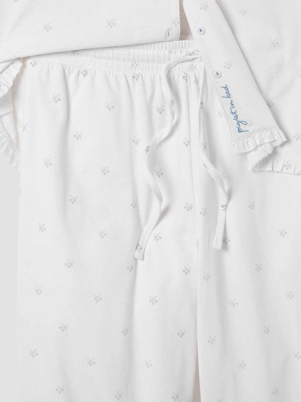 Close-up of a white pajama set with a small blue floral pattern, featuring a drawstring waist and a matching long-sleeve top with ruffled edges and blue script embroidery.