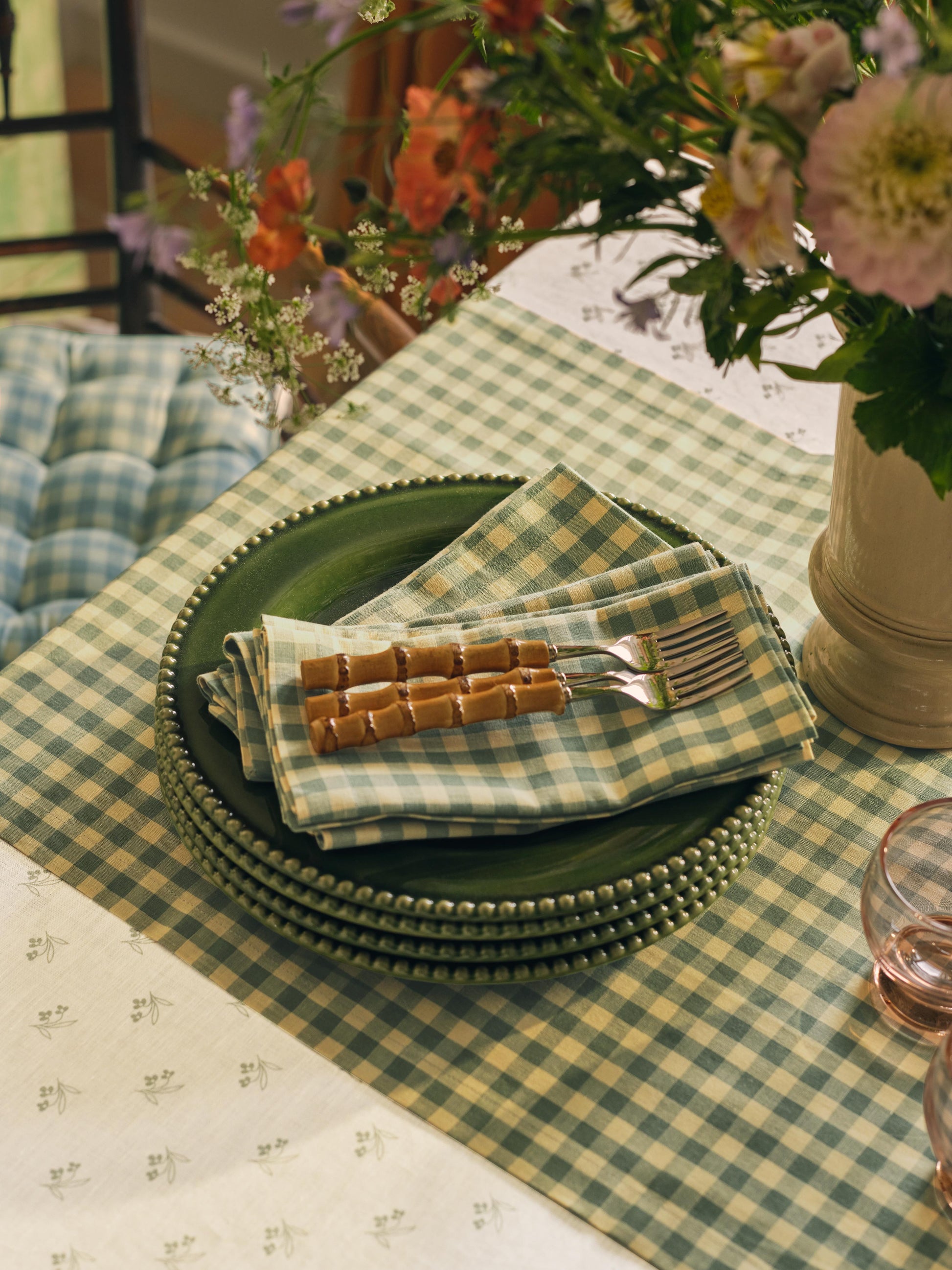 Green gingham table runner and white floral tablecloth with plates, napkins, and flowers on a table. 