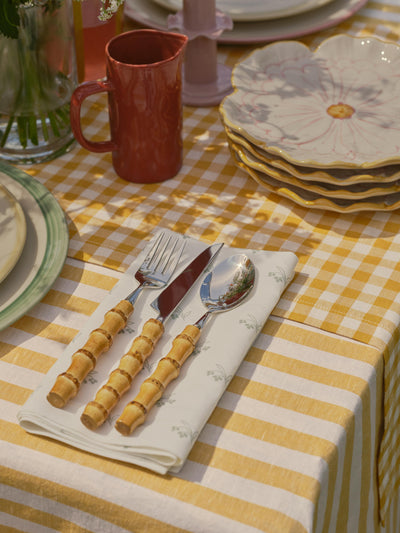 Table setting with cutlery on a white napkin with subtle green florals, on a yellow striped tablecloth.