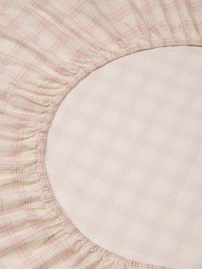 Close-up of a fitted sheet with a soft ruffled edge, featuring a light beige and white plaid pattern with thin pink and green lines.