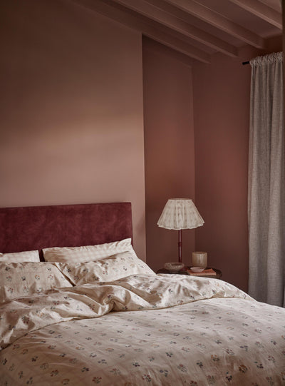 A cozy bedroom scene featuring a bed dressed in cream floral bedding and a dark burgundy velvet headboard, set against dusty rose walls with a bedside table, lamp, and curtain.