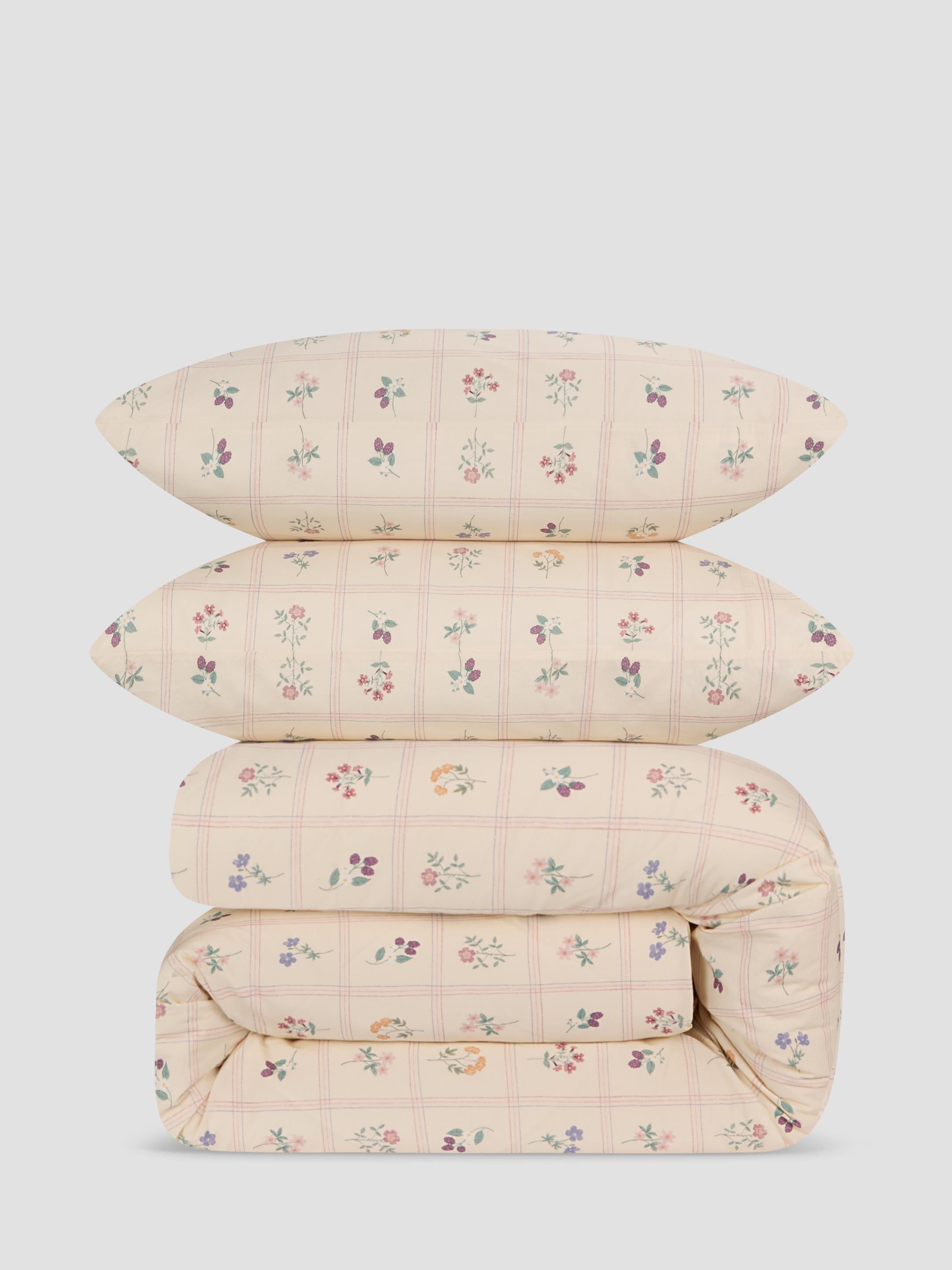 A stack of four bedding items, including two pillows and a folded duvet, in a soft cream color with a delicate pink grid pattern and small multicolored floral prints, set against a solid light gray background.
