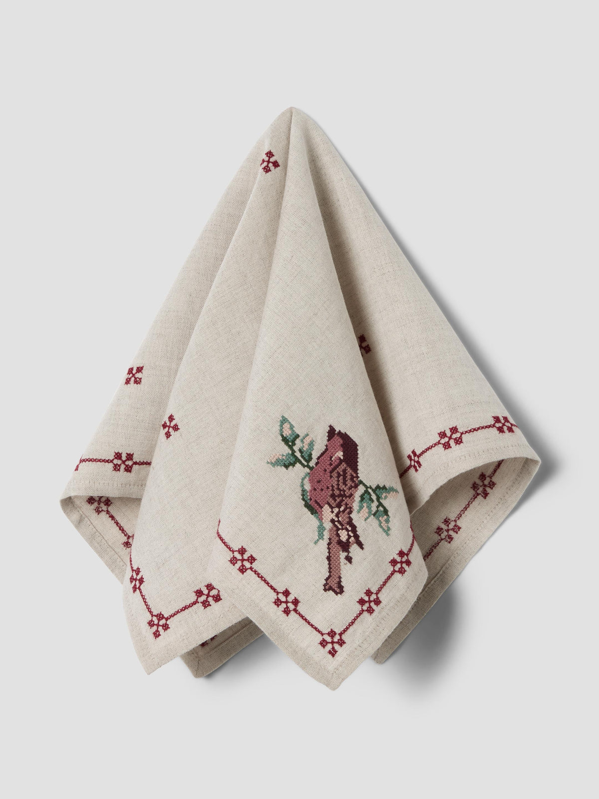 A folded beige linen napkin with embroidered red geometric borders and a central bird motif, displayed on a light gray background.