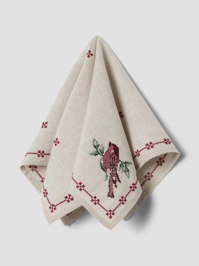 A folded beige linen napkin with embroidered red geometric borders and a central bird motif, displayed on a light gray background.
