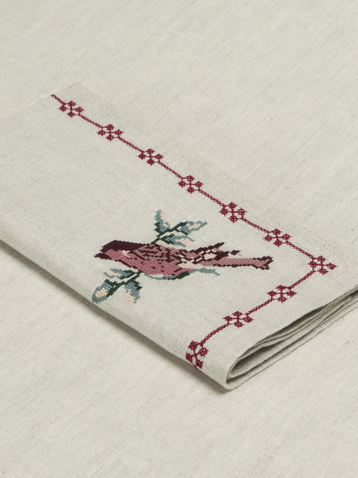 A folded cream-colored linen napkin featuring a detailed embroidered bird in pink and green, bordered by a red geometric pattern.