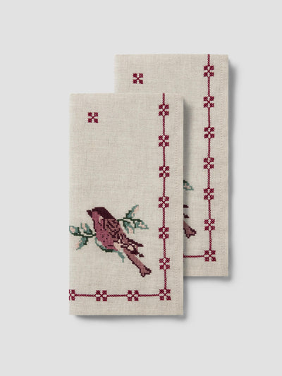Two cream-colored linen tea towels with embroidered bird and geometric border designs in burgundy and green, displayed against a light gray background.