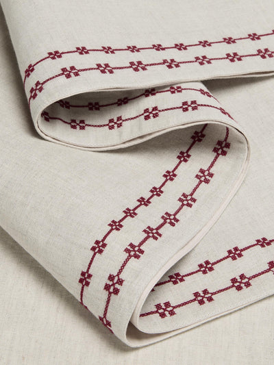 A close-up of a folded off-white linen tablecloth or napkin featuring a repeating dark red embroidered geometric pattern.