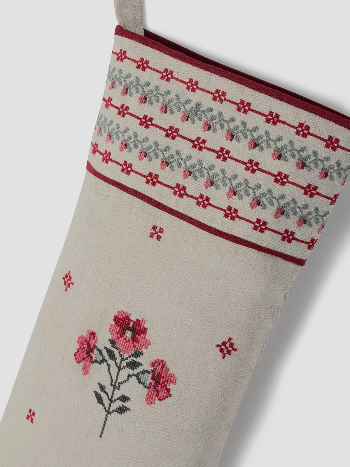A close-up of a cream-colored Christmas stocking with red and pink floral embroidery, a red and gray patterned cuff, and a hanging loop at the top.