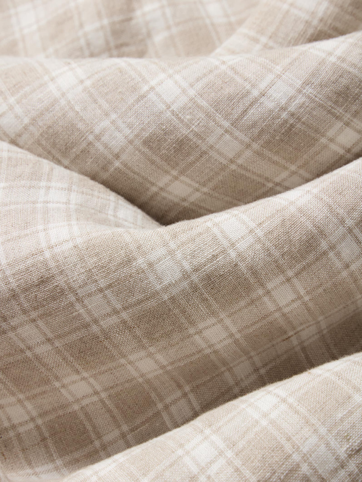 A close-up of draped fabric with a soft beige and off-white plaid pattern, showing the texture and folds of the material.