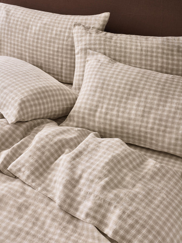 A close-up of a bed dressed in beige and brown gingham check bedding, including pillows and a duvet, with a dark brown headboard in the background.