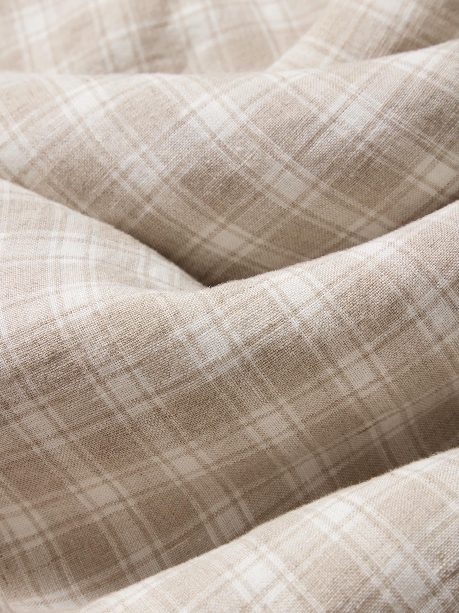 A close-up of draped fabric with a soft beige and off-white plaid pattern, showing the texture and folds of the material.