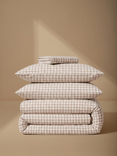 A stack of four folded bedding pieces, including two pillows and two folded blankets, in a beige and white gingham check pattern, set against a warm beige background with soft diagonal shadows.