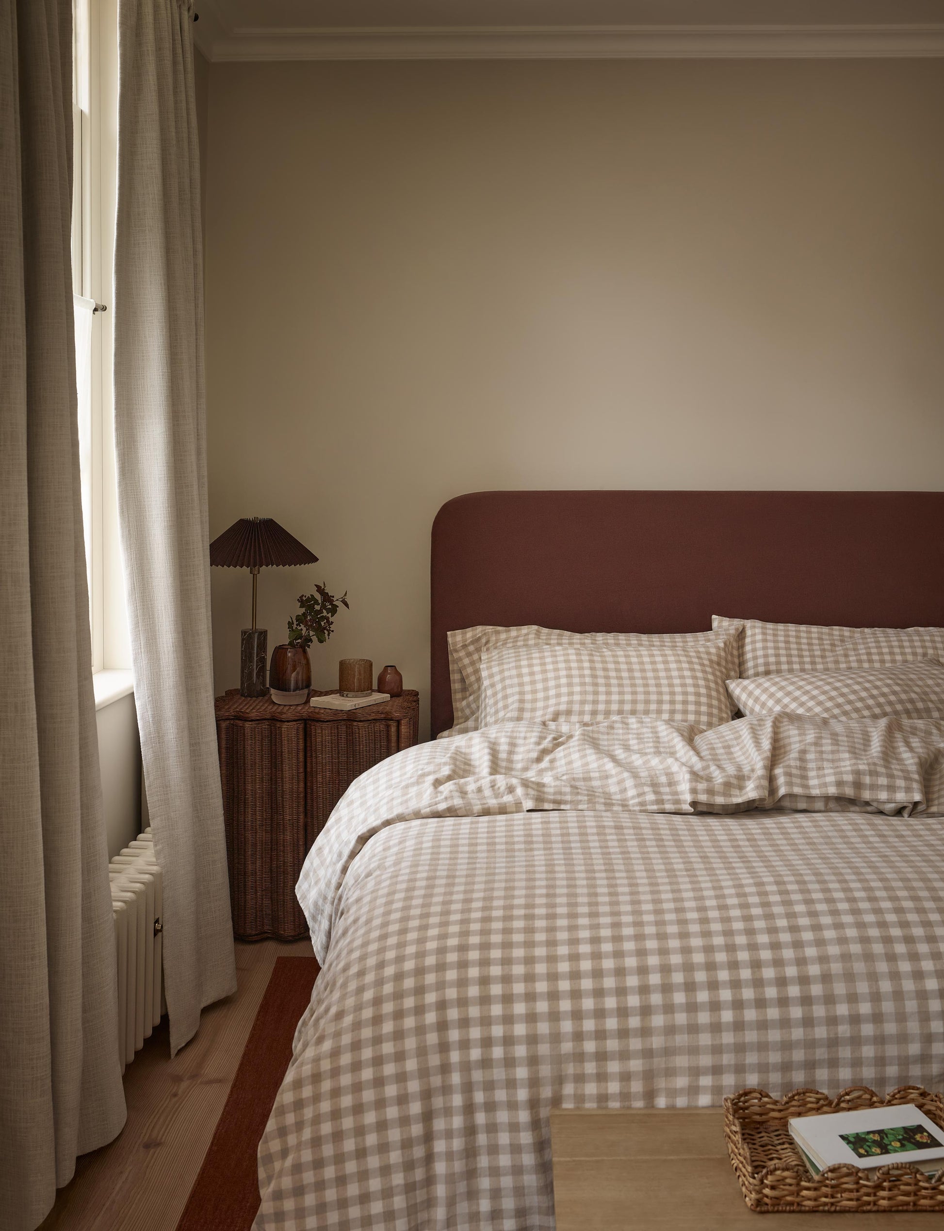A cozy bedroom scene featuring a bed dressed in beige and white gingham check bedding, set against a dark reddish-brown headboard. The room has warm beige walls, a wooden floor, a window with light-colored curtains, and a small bedside table with a lamp.