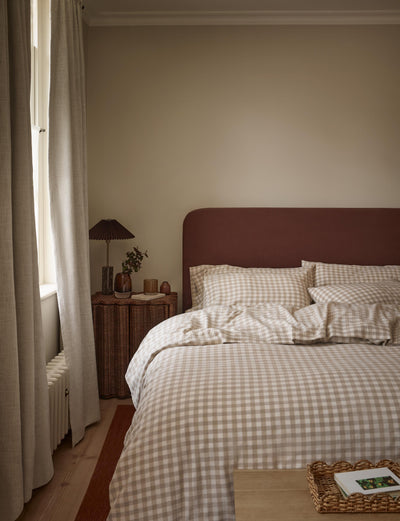 A cozy bedroom scene featuring a bed dressed in beige and white gingham check bedding, set against a dark reddish-brown headboard. The room has warm beige walls, a wooden floor, a window with light-colored curtains, and a small bedside table with a lamp.
