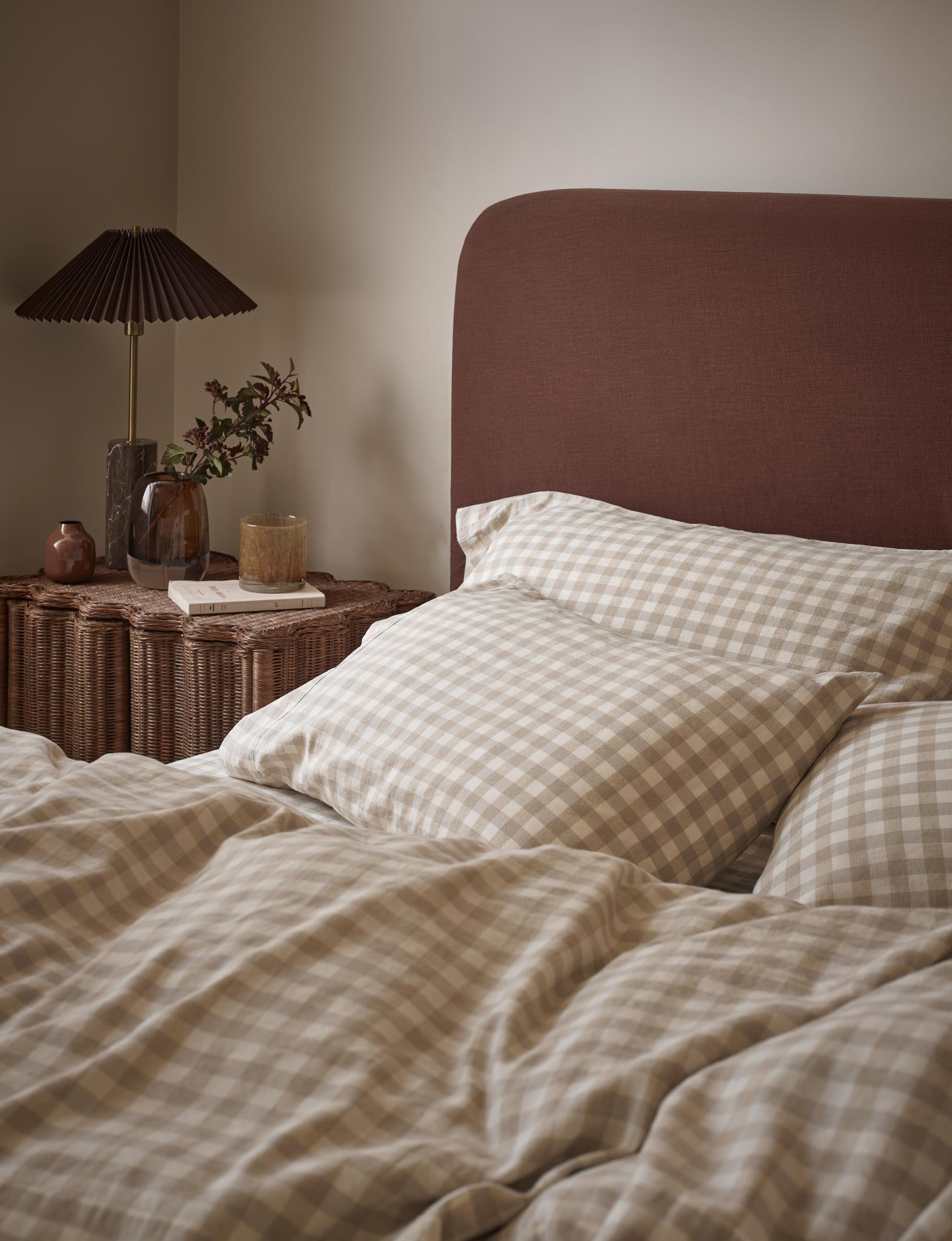 A cozy bedroom scene showing a bed made with beige and white gingham check bedding, including duvet cover and pillowcases, with a dark brown upholstered headboard. A wicker bedside table with a pleated black lamp and small plants is visible against a warm beige wall.
