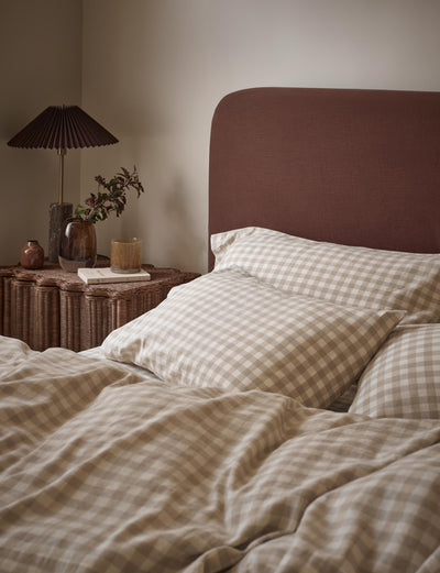 A cozy bedroom scene showing a bed made with beige and white gingham check bedding, including duvet cover and pillowcases, with a dark brown upholstered headboard. A wicker bedside table with a pleated black lamp and small plants is visible against a warm beige wall.