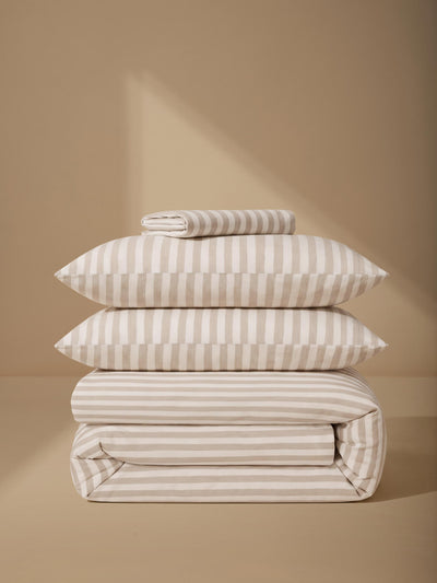 A stack of folded bedding, including pillows and a duvet cover, featuring a warm beige and off-white stripe pattern, set against a matching beige background with soft diagonal shadows.