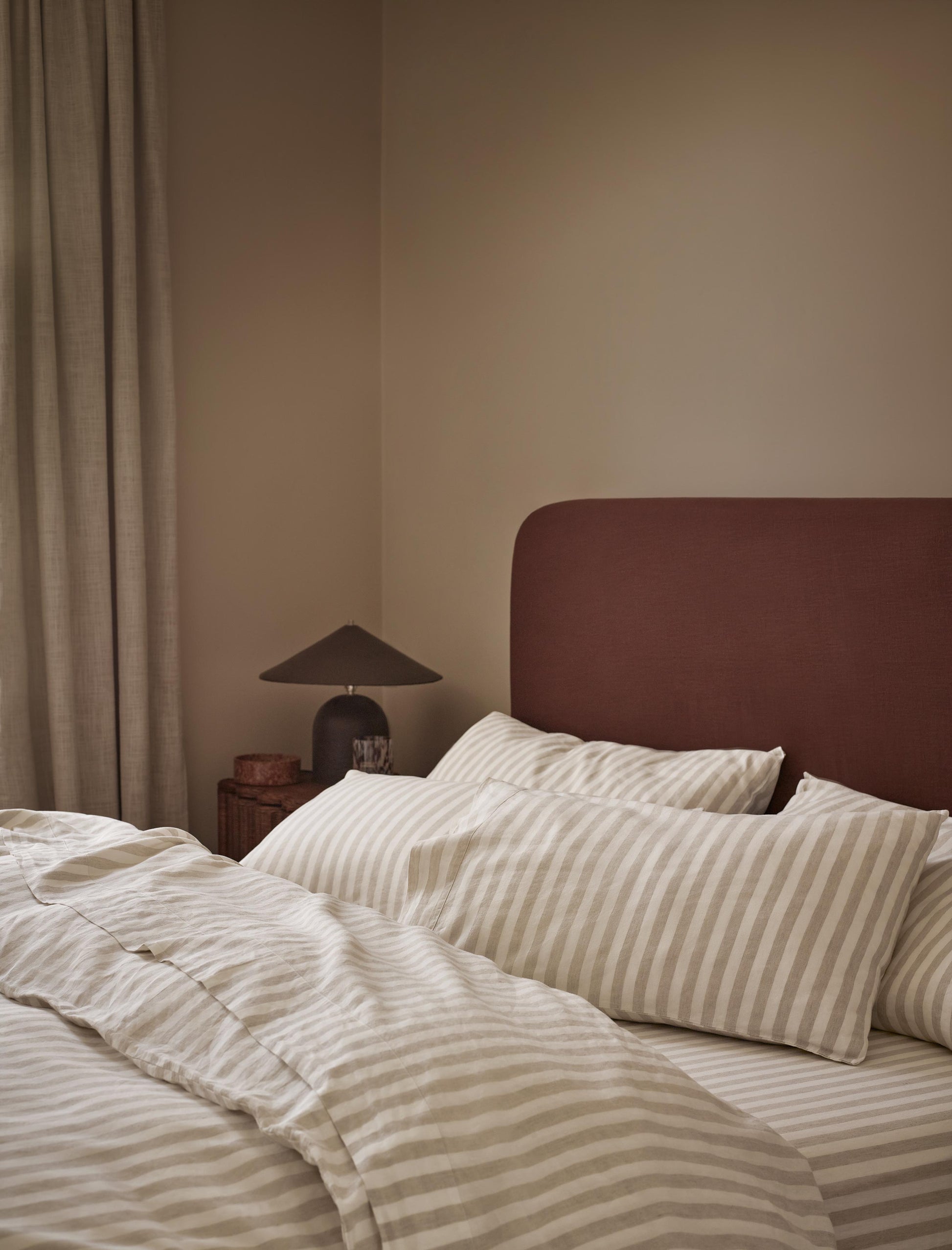 A cozy bedroom scene with a bed dressed in cream and beige striped bedding, a dark reddish-brown upholstered headboard, a bedside lamp, and neutral curtains against a warm beige wall.