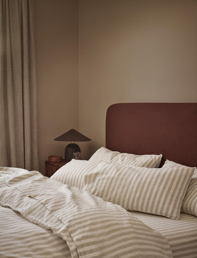 A cozy bedroom scene with a bed dressed in cream and beige striped bedding, a dark reddish-brown upholstered headboard, a bedside lamp, and neutral curtains against a warm beige wall.