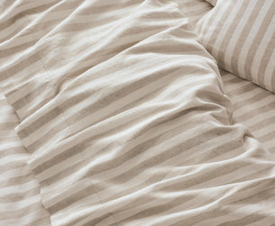 Close-up of striped bedding in warm beige and off-white tones, showing rumpled fabric and part of a matching pillow.