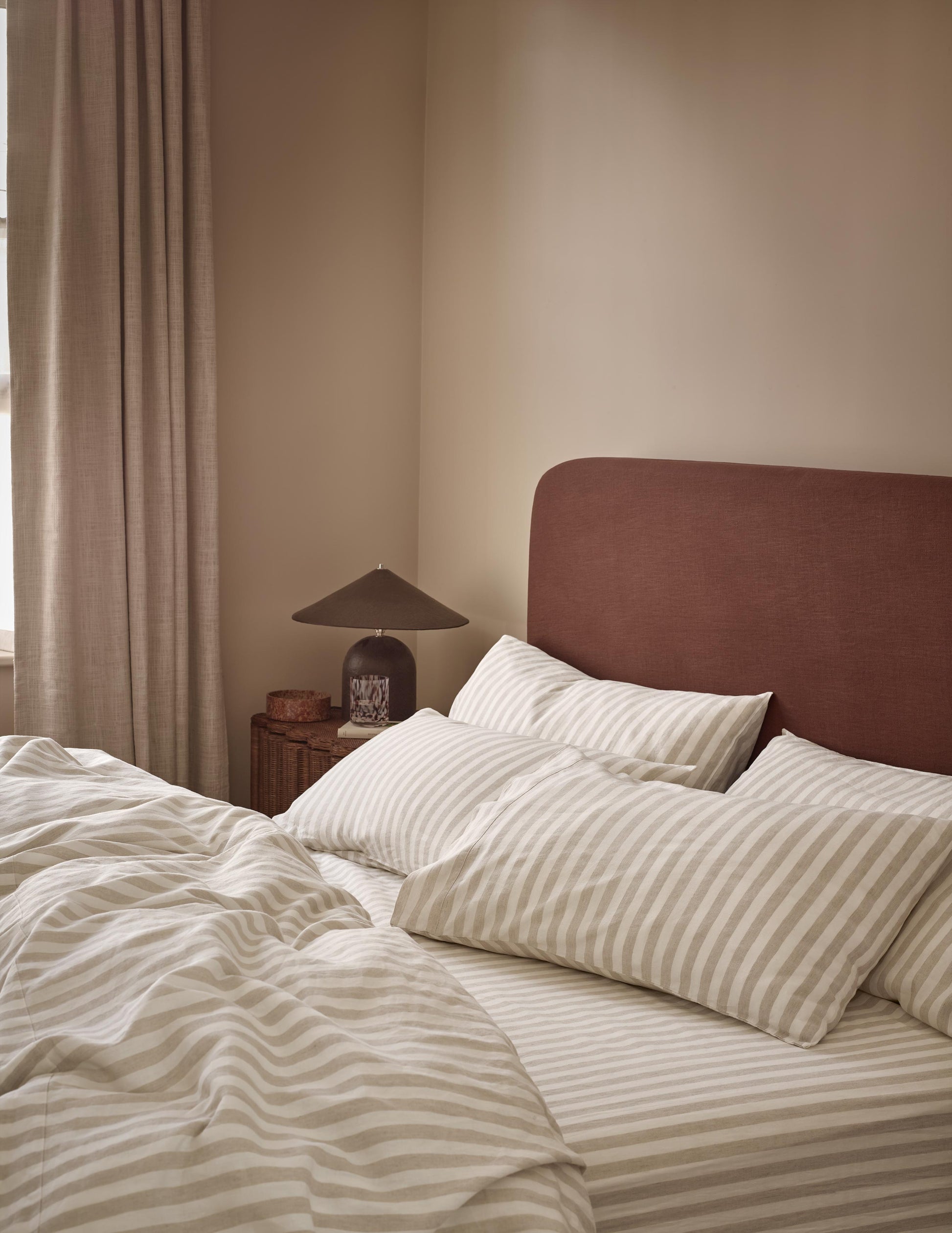 A styled bedroom scene featuring a bed with cream and beige striped bedding, a dark reddish-brown upholstered headboard, and a bedside table with a lamp against a warm beige wall.