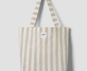 An oatmeal and white striped tote bag with a white label reading "Piglet in bed" on a light grey background.